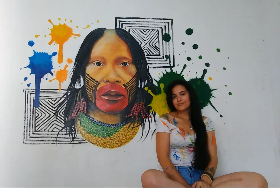 Cíntia Cris beside Ancestralidade em Cores, a mural with Indigenous visual motifs and vibrant graphic patterns.