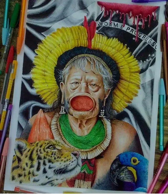 Ballpoint-pen portrait of Cacique Raoni Metuktire, a Mẽbêngôkre (Kayapó) leader, with a yellow feather headdress, the Brazilian flag, a jaguar and a macaw. Drawing by Cíntia Cris.